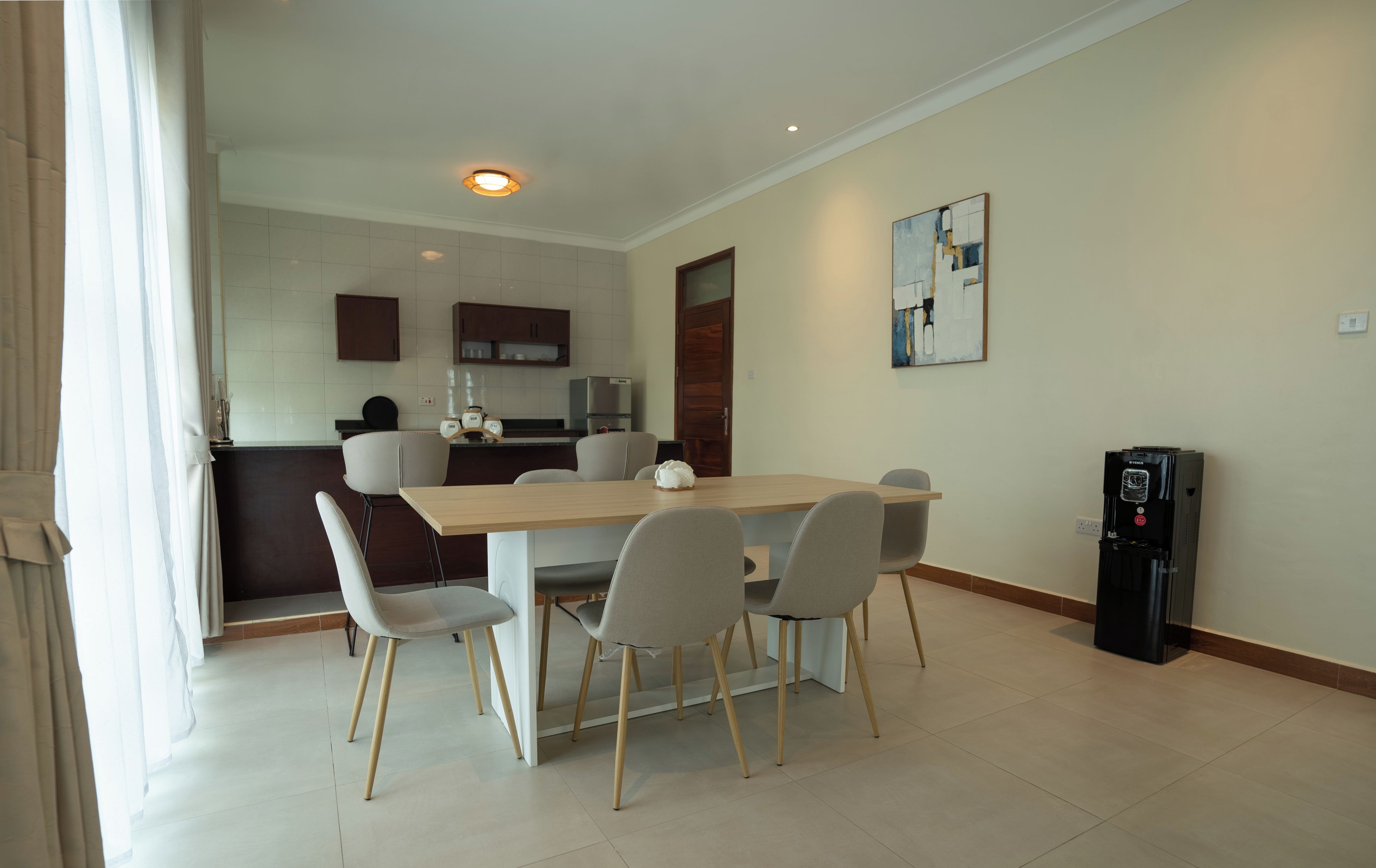 A dining area with a table and chairs, a kitchen in the background, and a water dispenser.