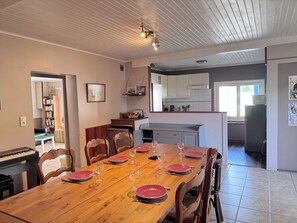 Ein Esszimmer mit einem Holztisch, gedeckt für sechs Personen, Küchenschränken, einem Kühlschrank und einem Klavier in der Ecke.