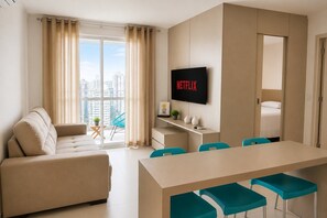 A modern hotel room with a sofa, a dining table with chairs, a TV, and a balcony with curtains.