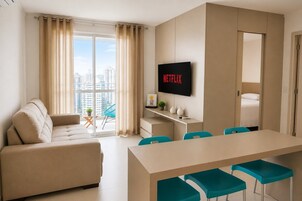 Quarto de hotel moderno com sofá, mesa de jantar com cadeiras, televisão e varanda com cortinas.