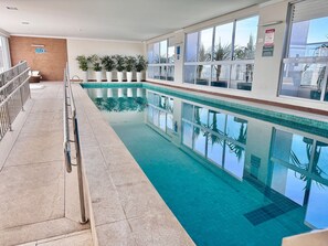 An indoor swimming pool with clear blue water, surrounded by windows, potted plants, and a tiled floor. - Walk Bueno (Goiânia)