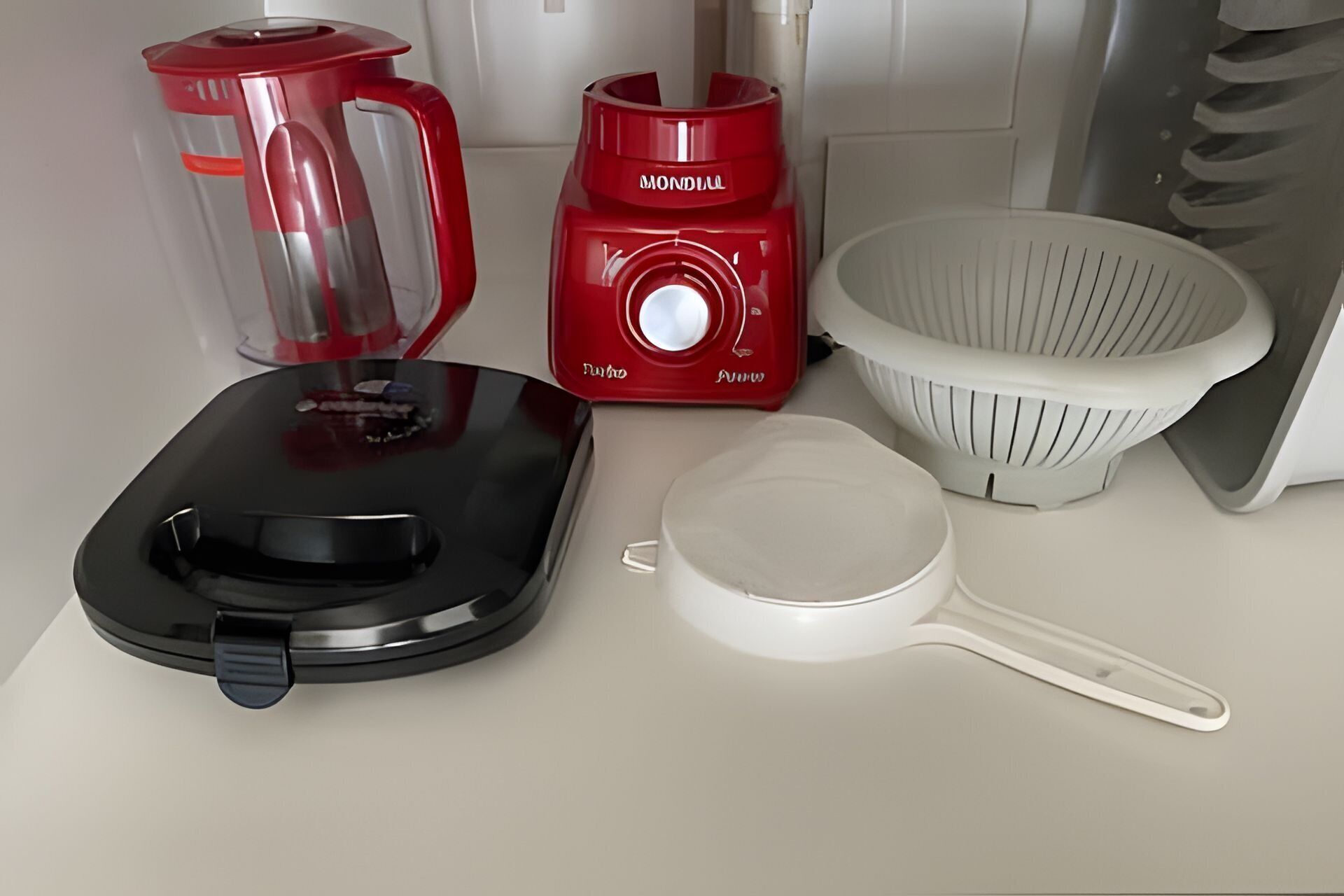 A kitchen area with a red blender, a black coffee maker, a white colander, and a white saucepan.