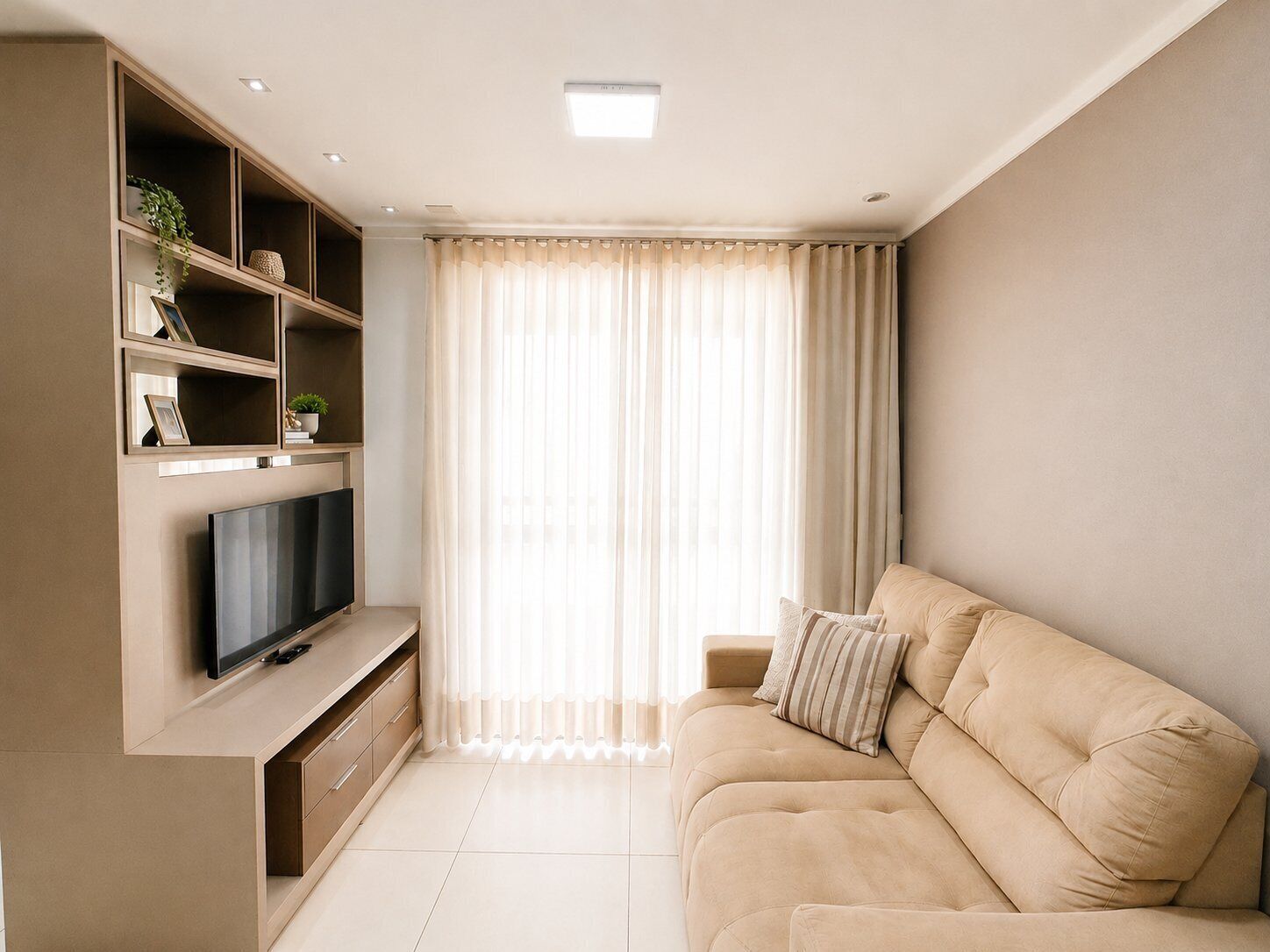 Ein modernes Wohnzimmer mit Fernseher, integrierten Regalen, einem beigefarbenen Sofa und einem großen Fenster mit leichten Vorhängen.