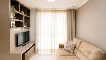 A modern living room with a flat-screen TV, built-in shelving, a beige sofa, and a large window with sheer curtains.