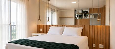 A modern hotel room with a large bed, a bedside table, a mirror, and a view of the city through the window.
