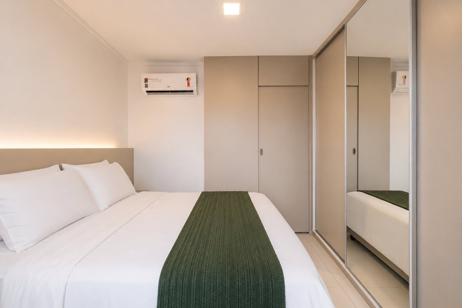 A hotel room with a bed, a green throw, a wardrobe, and an air conditioning unit.