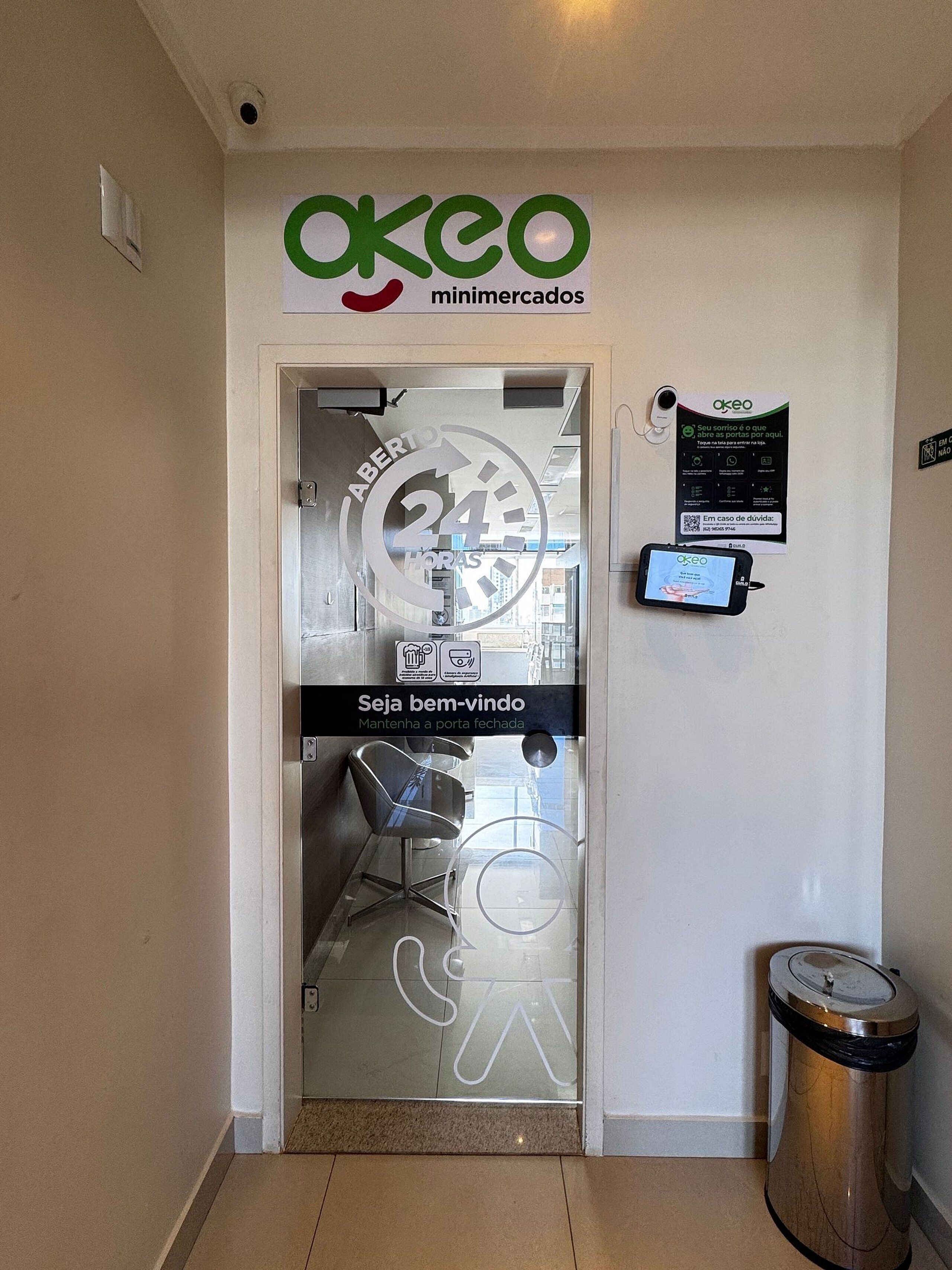 A door with a glass panel, a sign for 'OKEO minimercados', a 24-hour access sign, and a QR code for access.