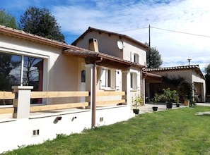 Une maison à deux étages dotée d’un balcon, d’une pergola et d’un jardin agrémenté de plantes en pot.