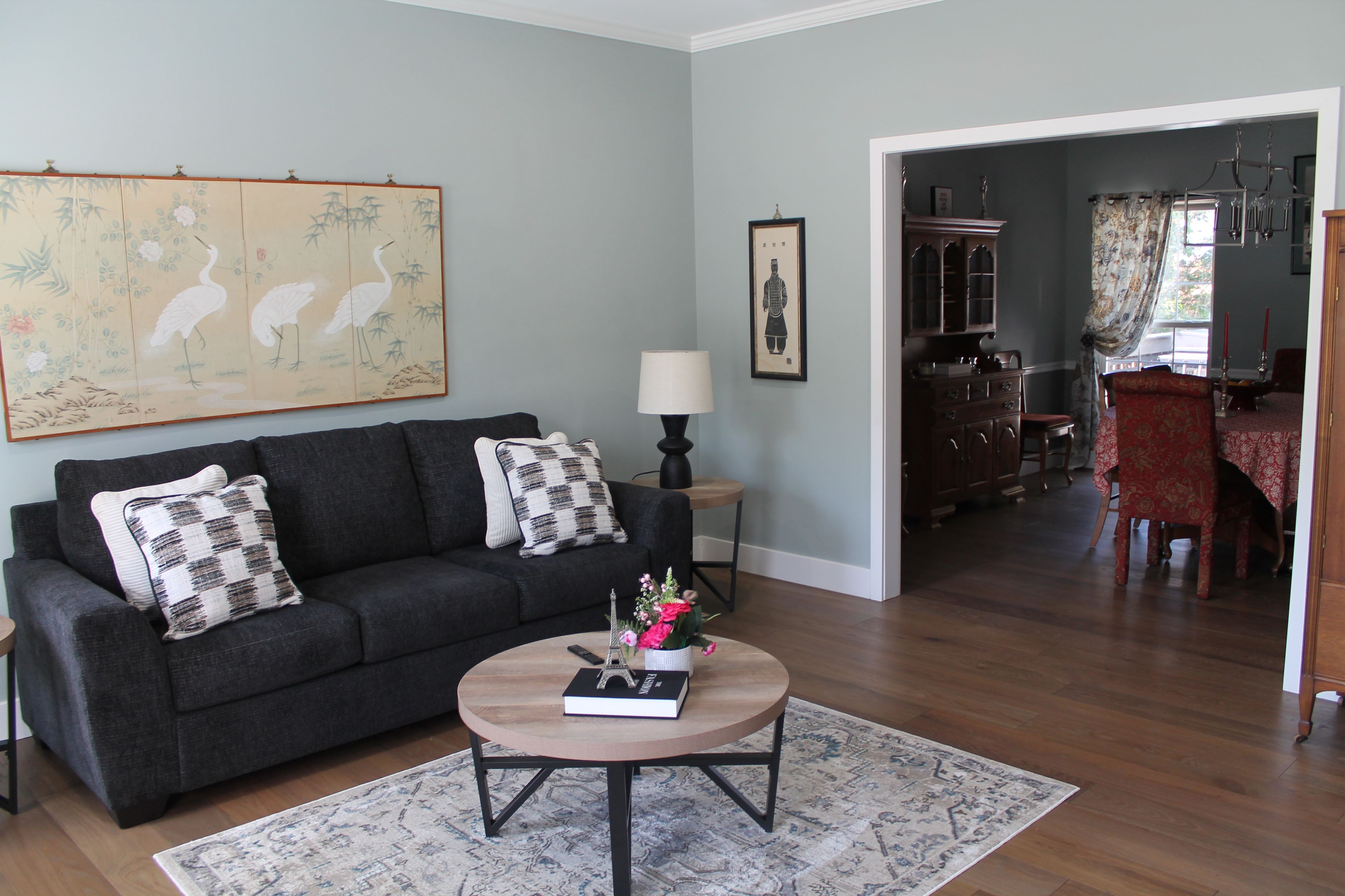 A living room with a dark grey sofa, a round coffee table with a book and flowers, a large painting of birds, and a dining area in the background.