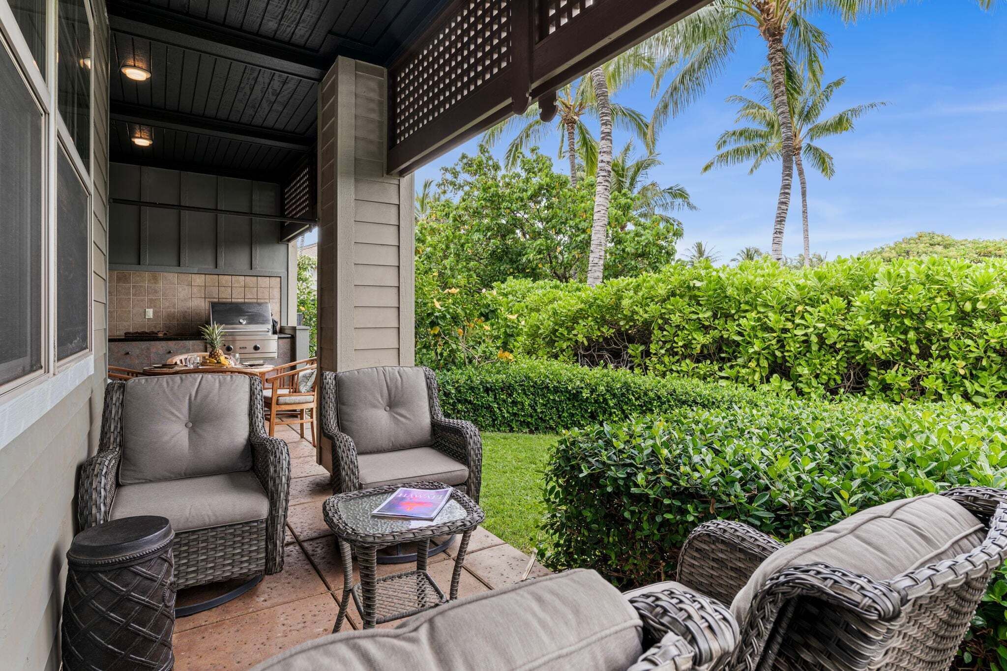 A covered outdoor seating area with wicker furniture, a grill, and a view of lush greenery and palm trees.