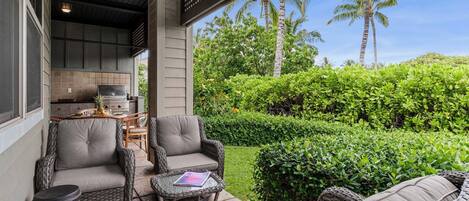 A covered outdoor seating area with wicker furniture, a grill, and a view of lush greenery and palm trees.