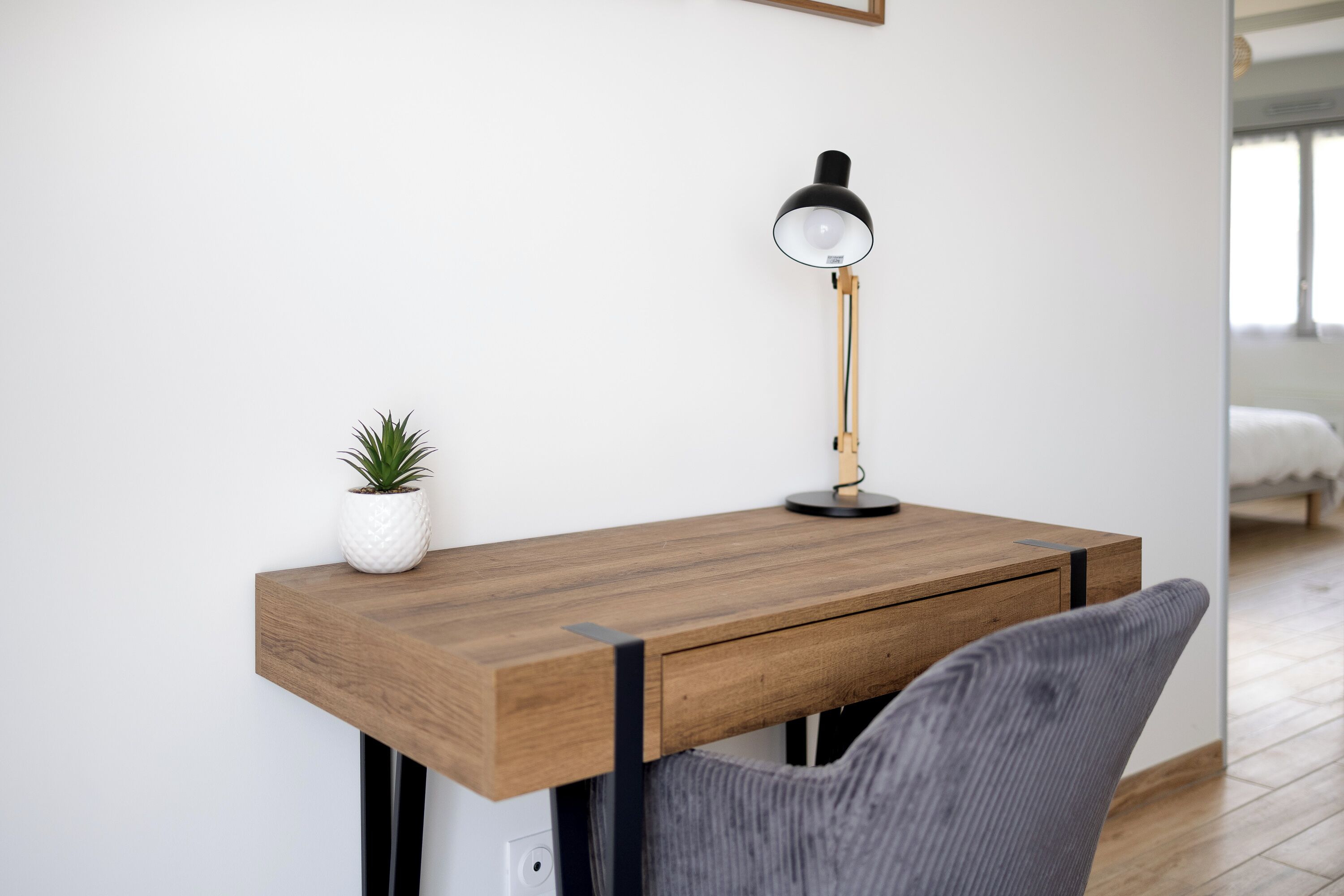 Un bureau en bois, une lampe, une petite plante en pot et une chaise grise.