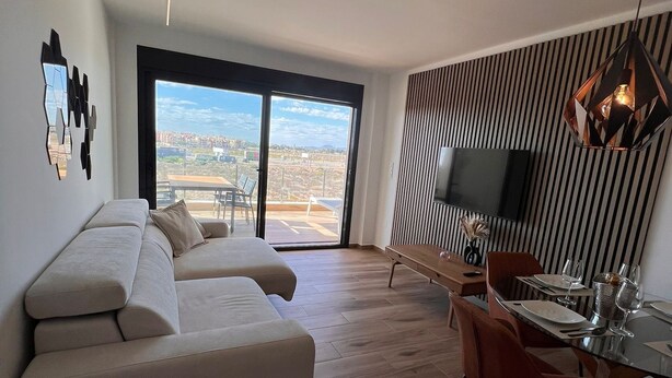 Un salon moderne comprenant un canapé, une table à manger et des chaises, un téléviseur fixé au mur et une grande fenêtre donnant sur la ville.