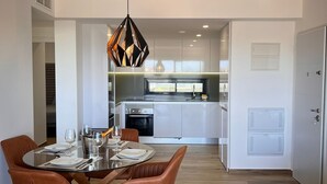 Une cuisine moderne avec des meubles blancs, un coin repas doté d’une table en verre et de chaises marron, ainsi qu’un luminaire suspendu.