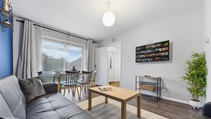 A modern living room with a sofa, dining table, chairs, a TV mounted on the wall, and a potted plant. - 4 Bed House Close To Gatwick Free Parking (West Sussex)