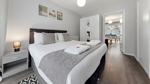 A modern hotel room with a large bed, bedside tables, a nightstand, a wardrobe, and a dining area visible through an open door. - 4 Bed House Close To Gatwick Free Parking (West Sussex)