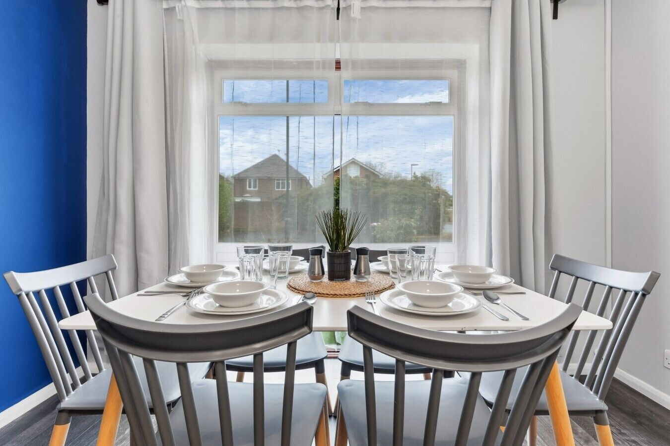 A dining area with a round table set for four, chairs, a potted plant, and a view of houses through a large window.