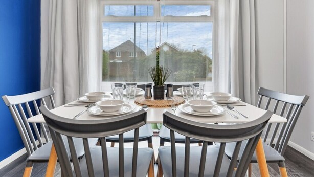 A dining area with a round table set for four, chairs, a potted plant, and a view of houses through a large window. - 4 Bed House Close To Gatwick Free Parking (West Sussex)