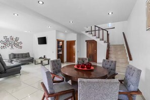 Ein geräumiges Wohnzimmer mit einem runden Holzesstisch, grau gepolsterten Sesseln, einem grauen Sofa, einem Flachbildfernseher und einer Treppe, die zu einer weiteren Ebene führt.