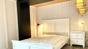Un dormitorio con cama blanca, una mesita de noche, una lámpara y un aplique de pared.