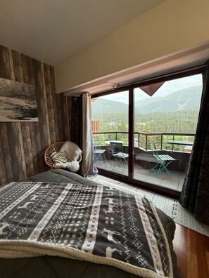Quarto com uma cama grande, uma cadeira de vime e uma varanda com vista para as montanhas.