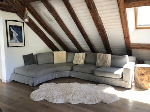 Ein Loft-Zimmer mit grauem Ecksofa, einem flauschigen weißen Teppich, Holzbalken, einem gerahmten Bild an der Wand und einem Holzhocker.