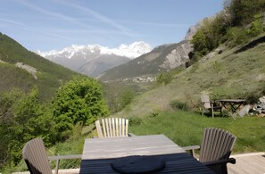 Una terraza de madera con dos sillas Adirondack y una mesa, con vista a un paisaje montañoso de picos nevados.