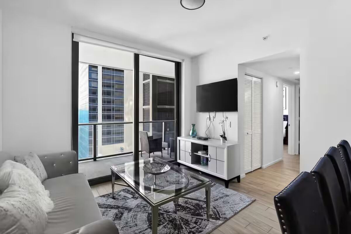 A modern living room with a glass coffee table, a sofa, a TV mounted on the wall, and a large window with blinds.