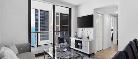 A modern living room with a glass coffee table, a sofa, a TV mounted on the wall, and a large window with blinds.