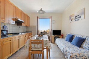 A kitchen with wooden cabinets, a dining table with chairs, a sofa, a TV, and a view through a doorway. - Apartment with Sea and Nature Views (Colle d'Orano)