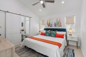 Une chambre à coucher comprenant un lit, des tables de chevet, un ventilateur de plafond, une fenêtre avec des stores et une porte.