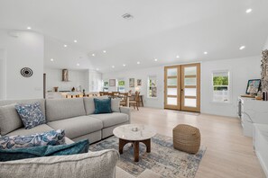 Ein geräumiger Wohnbereich mit Sofa, Couchtisch und Hocker, im Hintergrund eine Küche und eine Glastür zu einem Balkon.