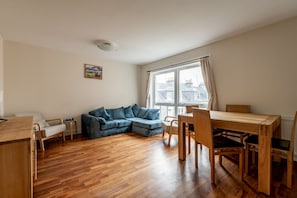 A room with a wooden dining table, a blue sofa, and a window with curtains. - Southfield Apartments (No 15) | St Andrews (St Andrews)
