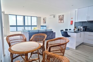 Eine moderne Küche mit weißen Schränken, ein Essbereich mit Korbstühlen und einem runden Tisch sowie eine Couch mit blauen Kissen.