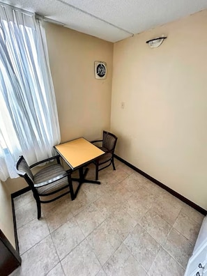 A small room with a table and two chairs, a window with curtains, a wall-mounted light, and a thermostat.