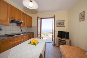 Una cucina compatta con mobili in legno, un tavolo da pranzo con un centro tavola di frutta, un televisore su un mobile basso e un balcone con vista.