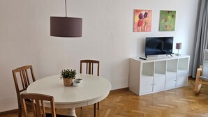A dining area with a round table, wooden chairs, a hanging light fixture, a white cabinet with a TV, and artwork on the wall.