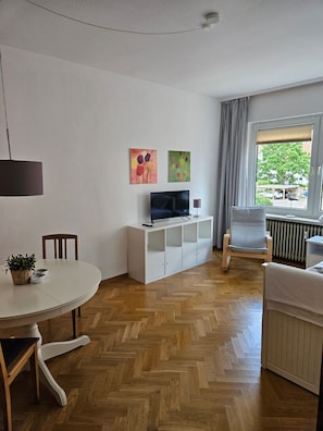 Ein modernes Wohnzimmer mit Fernseher, weißen Schränken, einem Esstisch und einem Sessel.