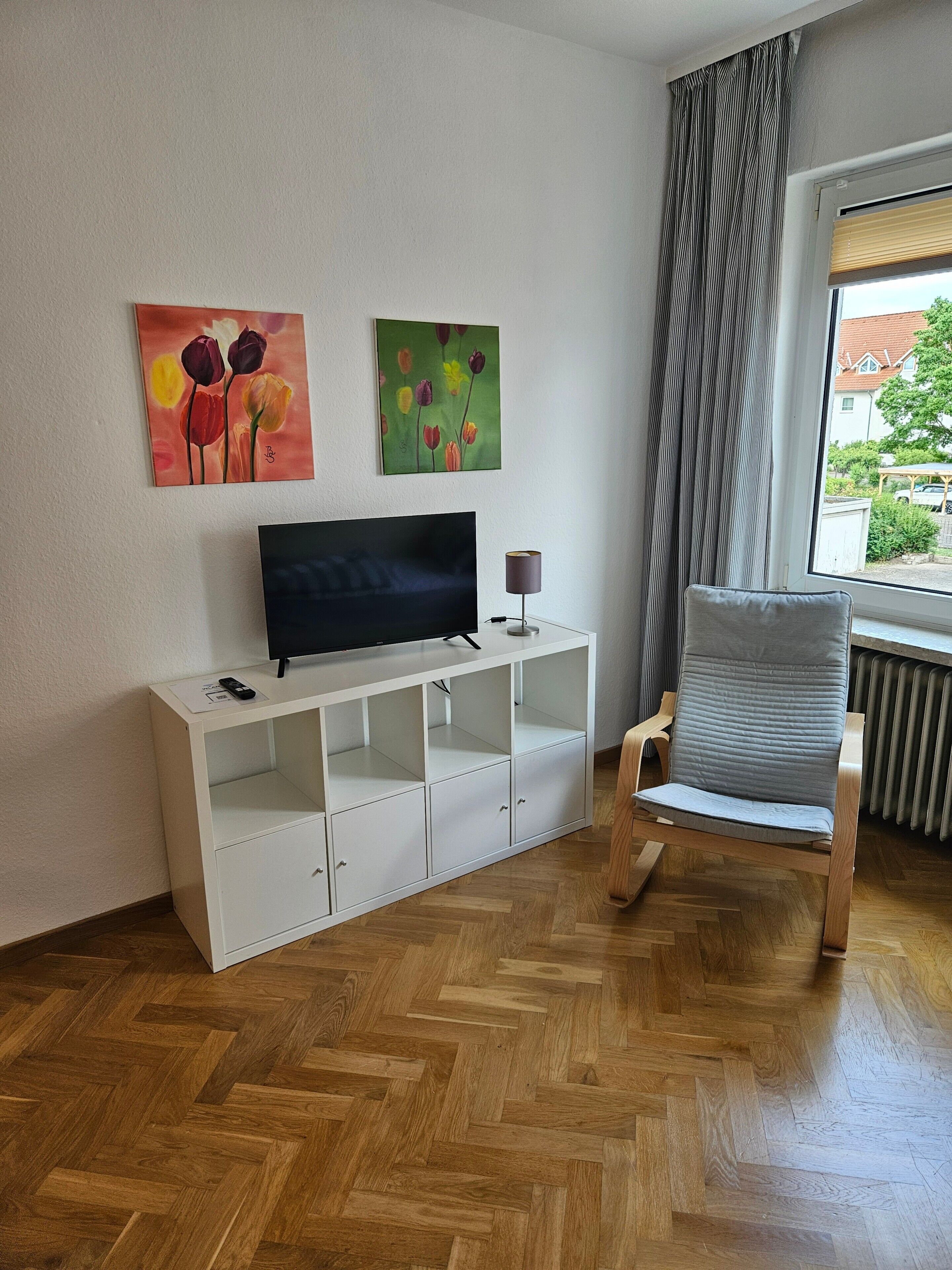 Una sala de estar con televisor, mueble de almacenamiento blanco, una silla de madera y dos cuadros en la pared.