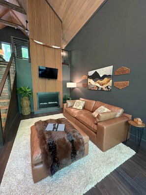 A modern living room with a leather sofa, a fur ottoman, a flat-screen TV, a fireplace, and decorative wall art. - Blackwood Retreat Cabin Hendersonville, NC (Henderson County)