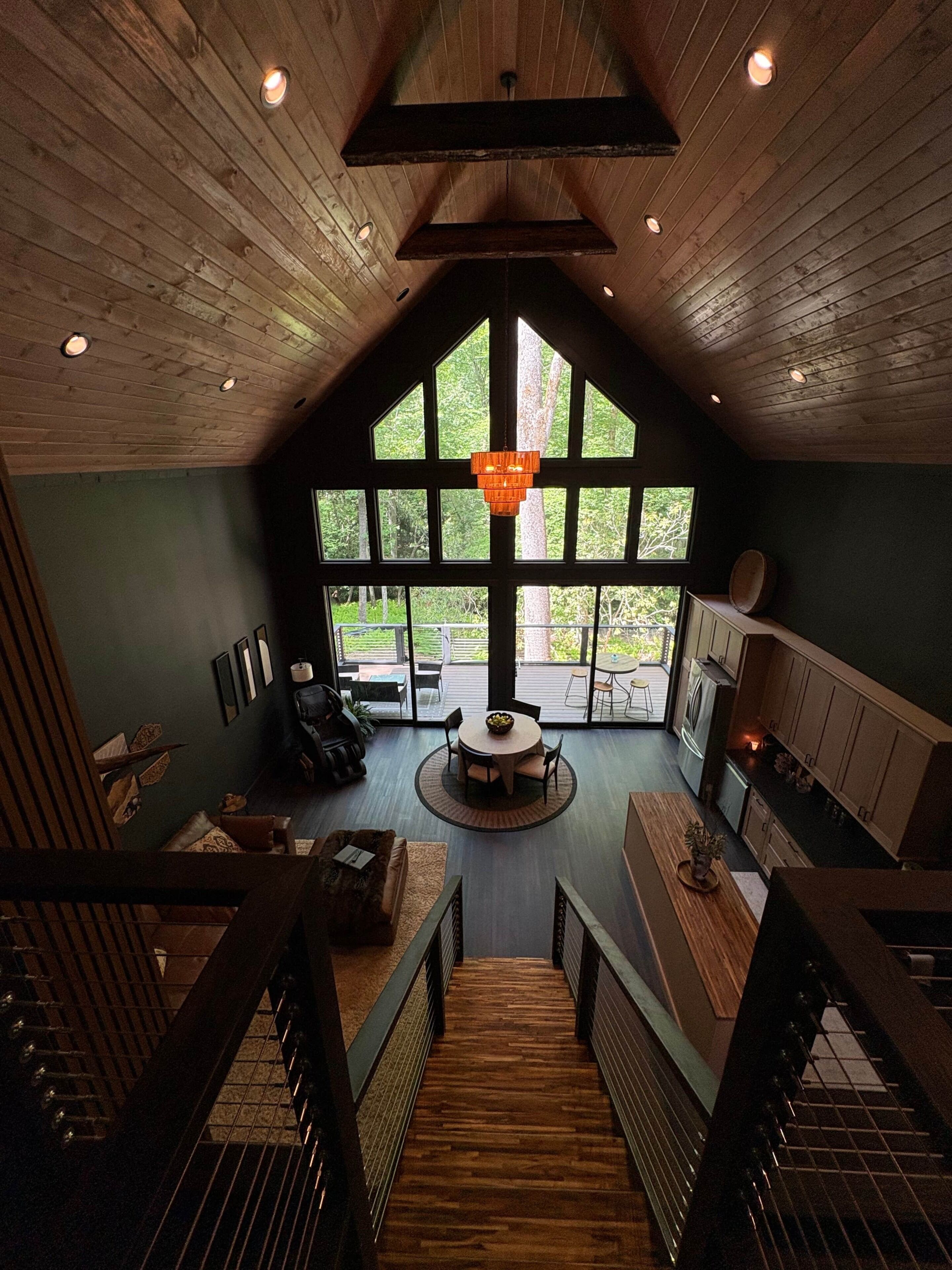 A spacious loft-style room with a high ceiling, large windows, a central chandelier, and a dining area with a round table.
