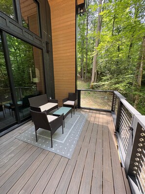 Eine Holzterrasse mit Gartenmöbeln, eine Glastür, die zum Innenbereich führt, und Blick auf ein bewaldetes Gebiet.