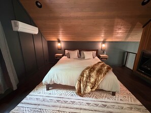 A bedroom with a large bed, two bedside tables, a wall-mounted air conditioner, and a fireplace. - Blackwood Retreat Cabin Hendersonville, NC (Henderson County)