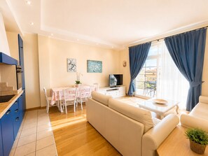 Ein Wohnzimmer mit Sofa, einem Esstisch mit Stühlen, einem Fernseher und einem Balkon mit blauen Vorhängen.