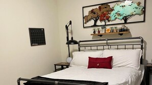 A single bed with a metal frame, white bedding, a red pillow, a bedside lamp, a wall-mounted shelf with decorative items, and a patterned rug. - Downtown 2 bedroom apartment. 20 minute walk to Sanford Stadium (Athens)