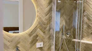 A modern bathroom with a herringbone tile wall, a round sink, a glass shower enclosure, and a mirror with a curved frame. - Villa Flavio-Panoramic Boutique Rooms  (Castelmola)