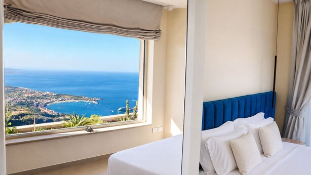 A bedroom with a large window offering a view of the sea and a coastal town. - Villa Flavio-Panoramic Boutique Rooms  (Castelmola)