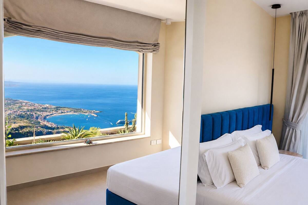 A bedroom with a large window offering a view of the sea and a coastal town.
