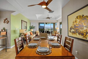 A dining area with a wooden table set for six, ceiling fans, a large framed artwork, and a view through the glass door. - Includes Beach Gear!  E305 Vista Waikoloa (Waikoloa)