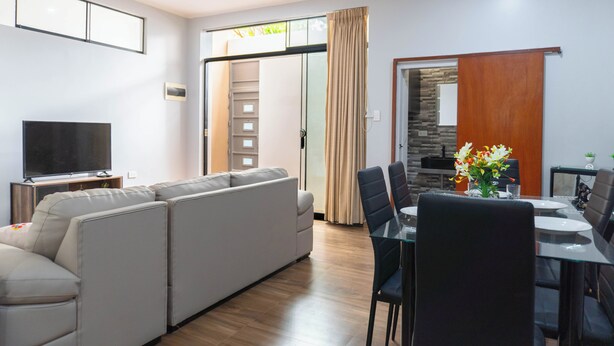 Una sala moderna con un sofá modular, una mesa de comedor con sillas, un televisor de pantalla plana y un ventanal corredero que da a una cocina.